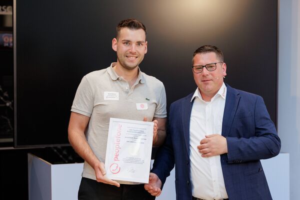 Zwei Personen stehen vor einem Flatscreen und halten eine Urkunde in die Kamera.