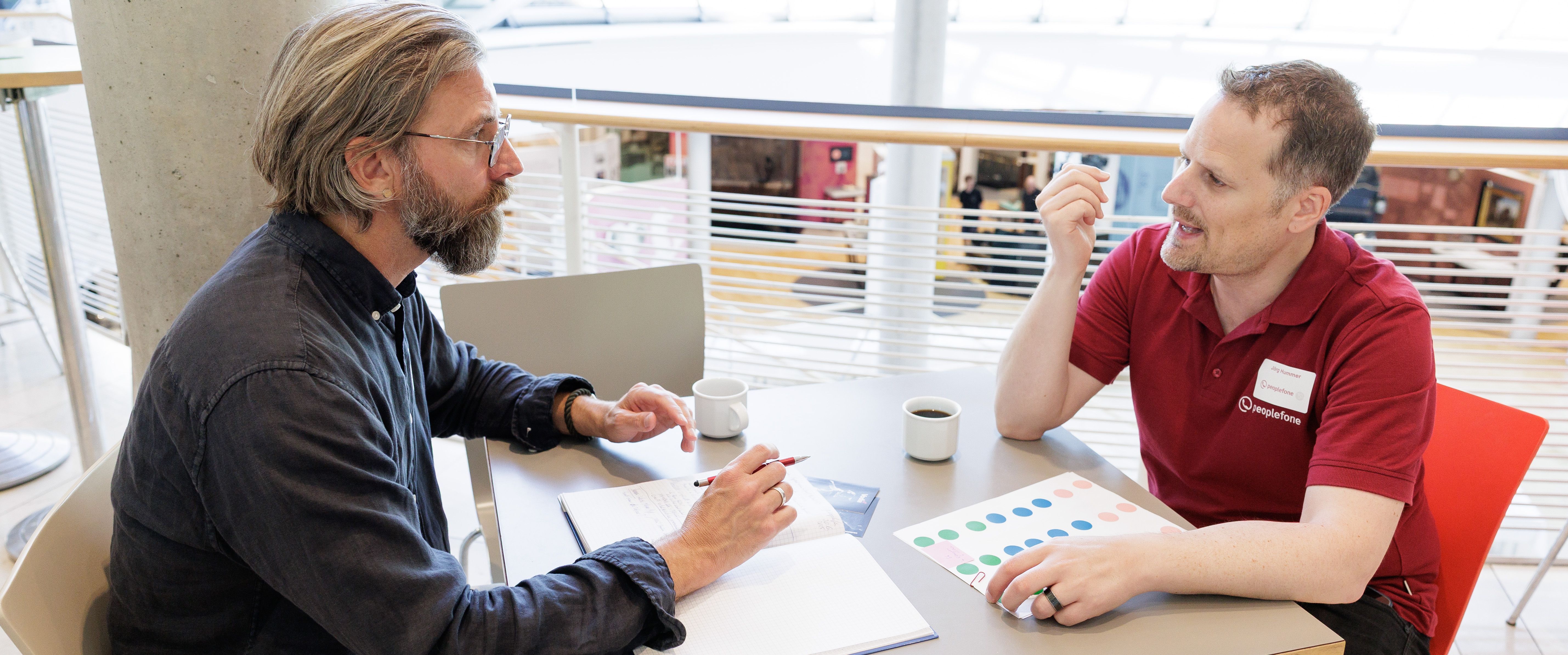 Zwei Personen sitzen am Tisch und unterhalten sich, während eine Person sich Notizen macht.