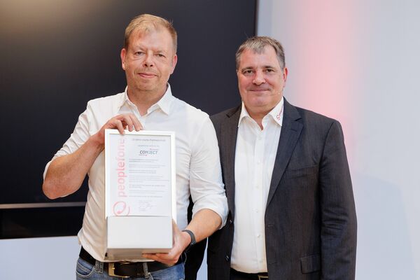 Zwei Personen stehen vor einem Flatscreen und halten eine Urkunde in die Kamera.