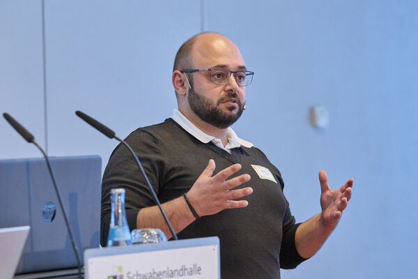 Eine Person steht auf der Bühne mit Mikrofon und hält einen Vortrag.