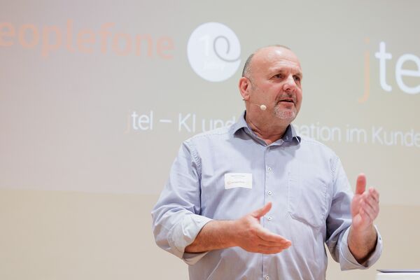 Eine Person steht an einem Stehtisch und hält einen Vortrag vor einer Leinwand.