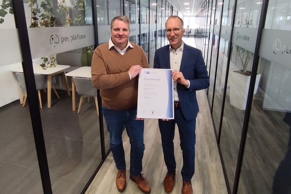 Zwei Personen stehen in einem Flur mit Glaswänden und halten eine Urkunde in die Kamera.