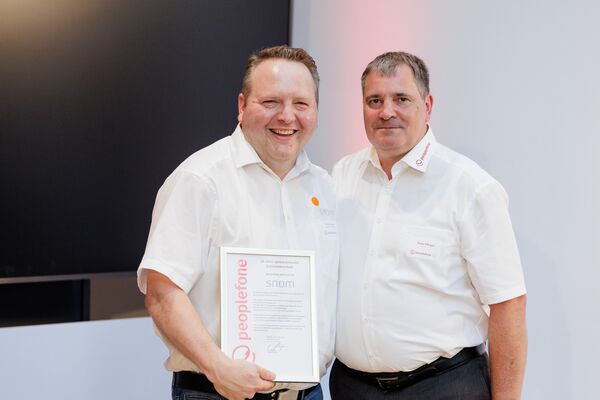 Zwei Personen stehen vor einem Flatscreen und halten eine Urkunde in die Kamera.