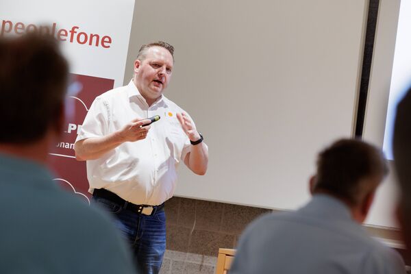 Eine Person steht vor einem Banner und hält vor einer Gruppe einen Vortrag.