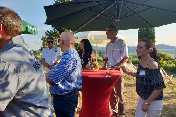 Personen an Stehtischen hören einem Vortrag zu. Sie stehen auf einer Wiese.