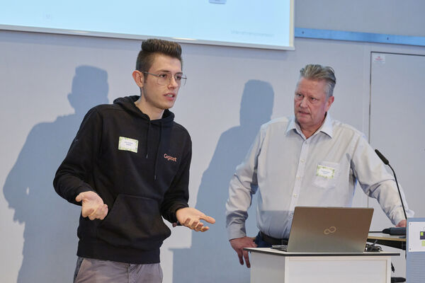 Zwei Personen stehen auf der Bühne mit Mikrofon und halten einen Vortrag.