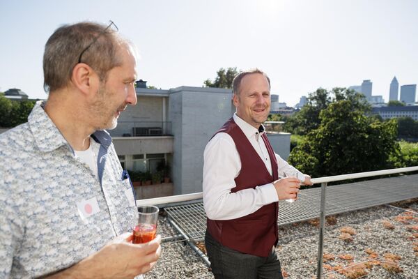 Zwei Personen stehen auf einer Dachterrasse. Im Hintergrund ist die Skyline von Frankfurt zu sehen.