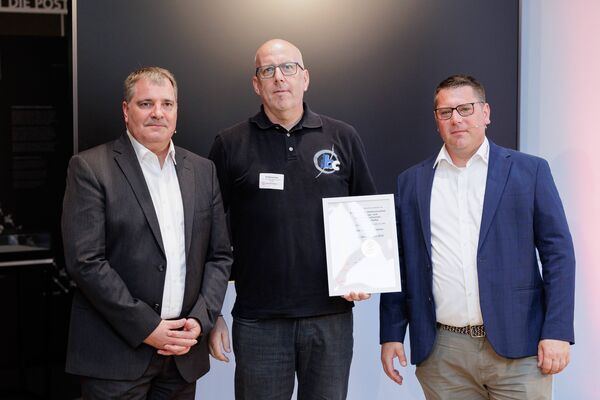 Drei Personen stehen vor einem Flatscreen und halten eine Urkunde in die Kamera.