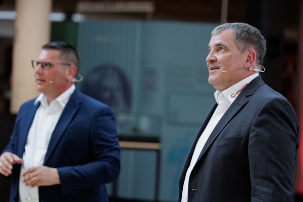 Zwei Personen stehen mit Headsets vor einer Wand und sprechen mit dem Publikum.