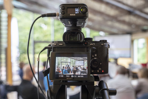 Blick durch eine Fotokamera in einen Vortragsraum mit sitzenden Personen.