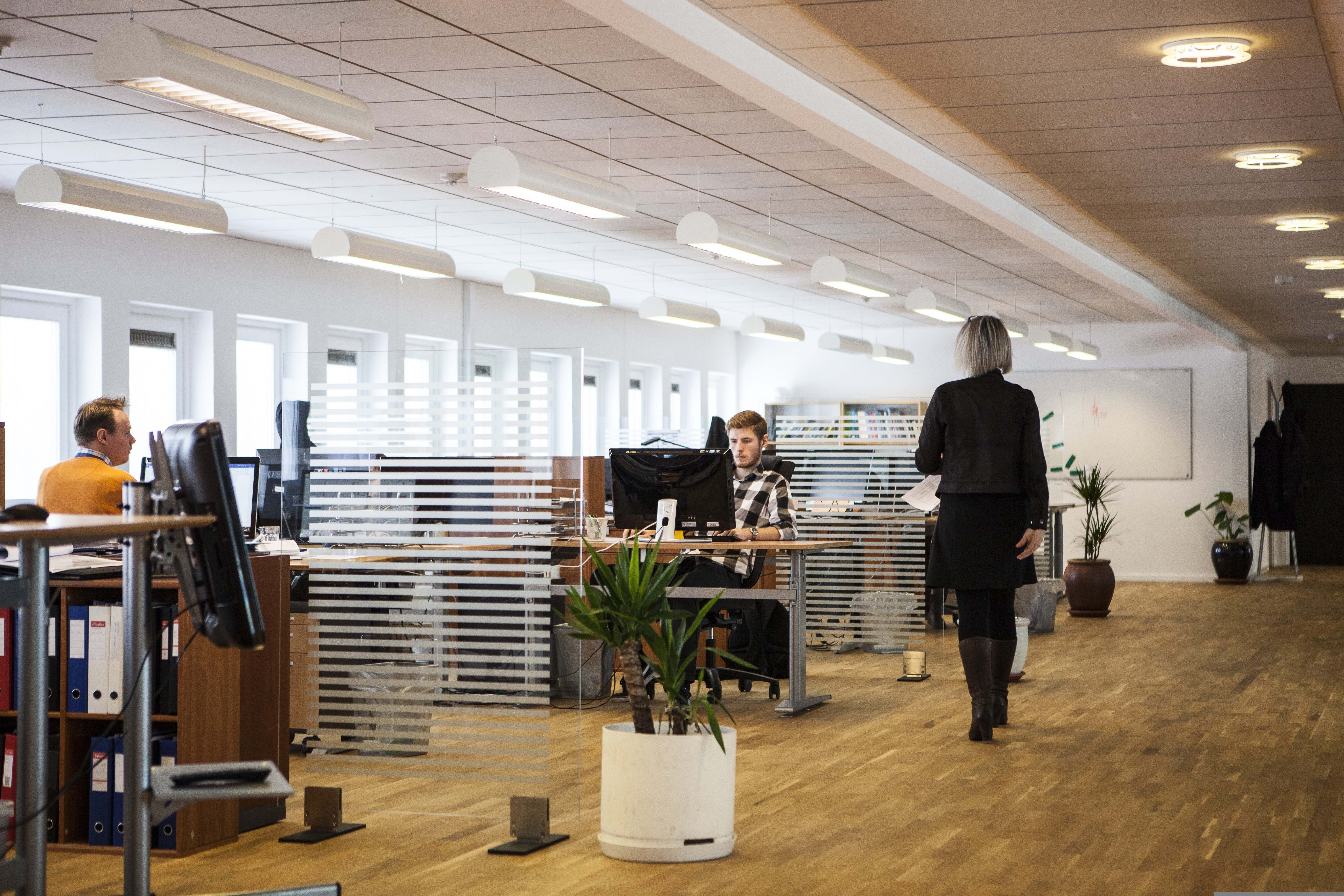 Büro mit mehreren Schreibtischen und Personen an einem langen Gang.