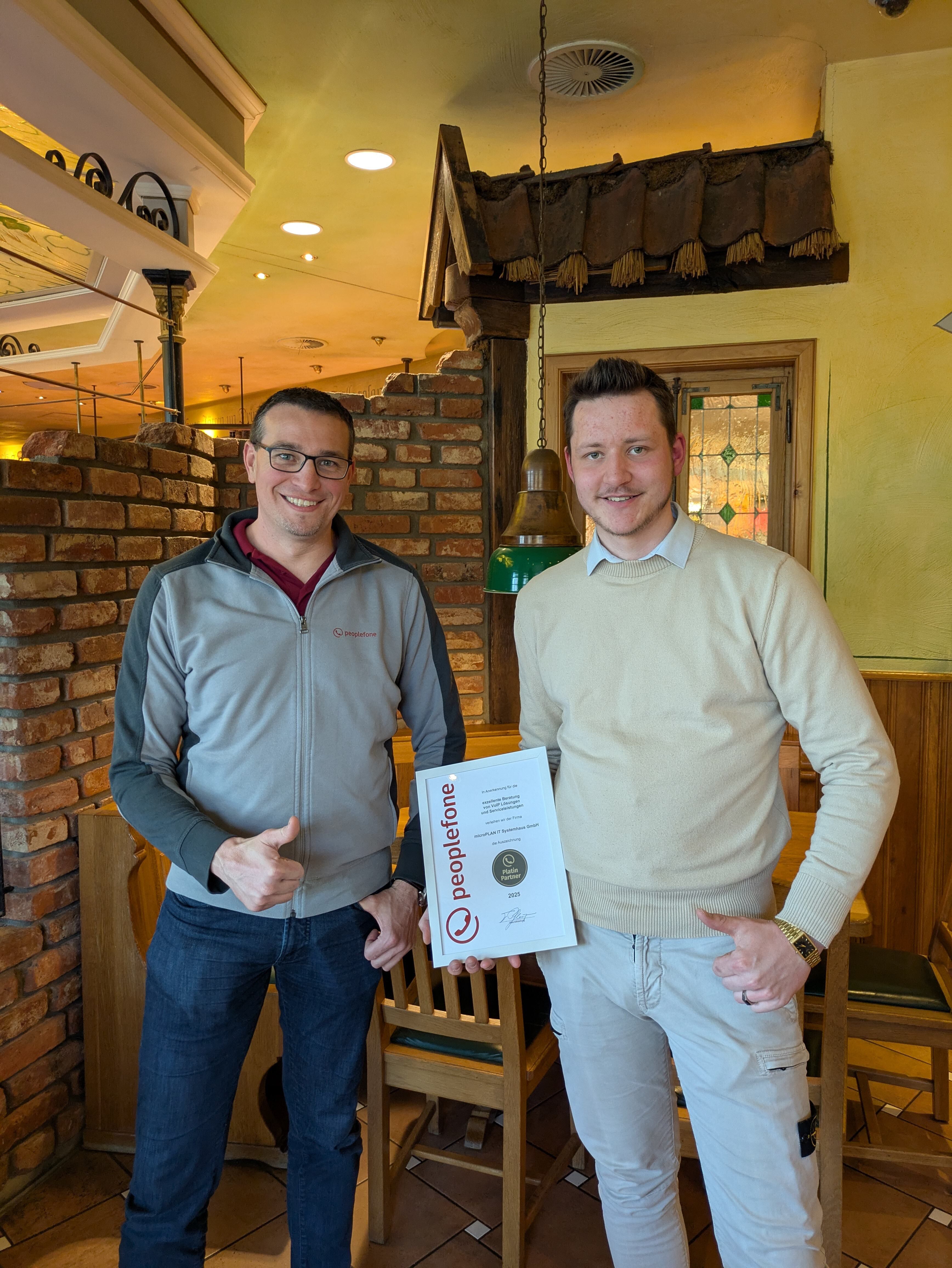 Zwei Personen stehen in einem Restaurant und halten eine Urkunde in die Kamera.